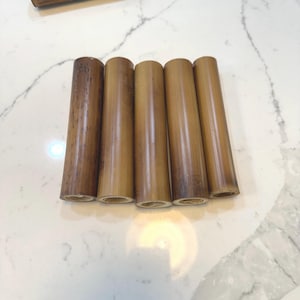 May include: Five natural brown bamboo tubes of varying lengths, arranged horizontally on a white marble surface.