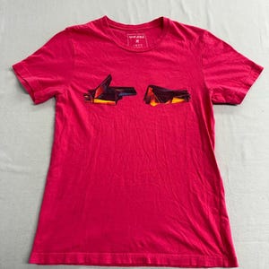 Puede incluir: Una camiseta rosa brillante con cuello redondo y mangas cortas. La camiseta presenta un diseño gráfico en la parte delantera, con el texto "RUN THE JEWELS" en la parte superior. El gráfico es un diseño abstracto y colorido.