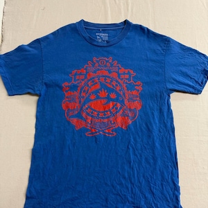 May include: A blue short-sleeve t-shirt featuring a large, intricate red graphic print on the chest. The design includes a central dolphin silhouette, stylized leaf shapes, and decorative patterns with text in both English and Asian characters.