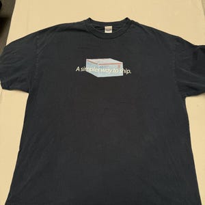 May include: A black short-sleeved t-shirt featuring a graphic of a white shipping box with red and blue accents. Below the box, the text reads, A simpler way to ship. The shirt has a crew neckline and a standard fit, presented on a flat surface.
