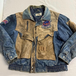 May include: A multi-textured jacket featuring a combination of blue denim and tan leather panels. The garment includes a fold-down collar, a front zipper closure, two large flap pockets, a vertical zippered chest pocket, and embroidered patches.