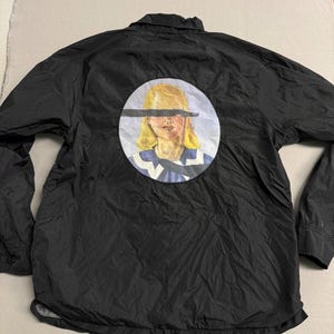 May include: Black nylon jacket with a button-up front and long sleeves. A circular graphic on the back features a portrait of a person with a black bar over the eyes. The person has blonde hair and is wearing a blue and white striped shirt.