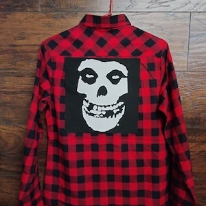 May include: Red and black plaid flannel shirt with a white skull graphic on the back.