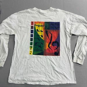 May include: White long-sleeve shirt featuring a colorful graphic of musical notes and a treble clef. The design incorporates red, yellow, green, and blue hues. The shirt has a crew neck and ribbed cuffs.