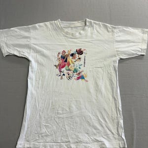 May include: White t-shirt featuring a colorful graphic print. The graphic depicts several figures in motion, with a mix of vibrant colors. The shirt has short sleeves and a crew neck, suitable for casual wear.