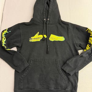 Puede incluir: Sudadera con capucha negra con bolsillo de canguro y capucha con cordón. La sudadera con capucha presenta gráficos amarillos y verdes en las mangas y el pecho. Los gráficos parecen ser formas abstractas y texto.