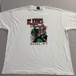 May include: White t-shirt featuring a graphic for St. John's Festa Italiana in Olean, NY. The design includes the text "St. John's Festa Italiana" in red and green, along with an illustration of the Leaning Tower of Pisa and a person.