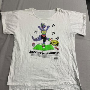 May include: White t-shirt featuring a cartoon dinosaur in workout attire, dancing with musical notes and a boombox. The shirt has the text "Jazzercisasaurus" with a definition below the title. A vintage, retro-themed design.