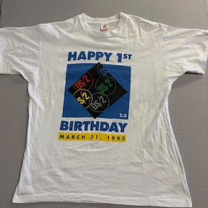 Op de afbeelding: Wit t-shirt met een grafisch ontwerp ter viering van de 1e verjaardag van OS/2. Het ontwerp bevat de tekst "HAPPY 1ST BIRTHDAY MARCH 31, 1993" in blauw en geel, met OS/2 logo's in een ruitvorm.