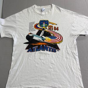 以下が含まれることがあります： 白いTシャツ。スペースシャトル・アトランティスのグラフィック、テキスト「STS 84」と「アトランティス」が特徴です。デザインには、様式化されたアメリカ国旗と宇宙をテーマにしたエンブレムが含まれています。Tシャツは半袖で、クルーネックです。