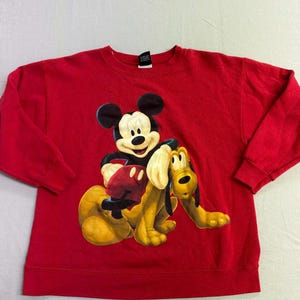 May include: A red sweatshirt featuring a graphic of Mickey Mouse and Pluto. Mickey is wearing a black jacket and red gloves, while Pluto is yellow. The sweatshirt is a solid red color.