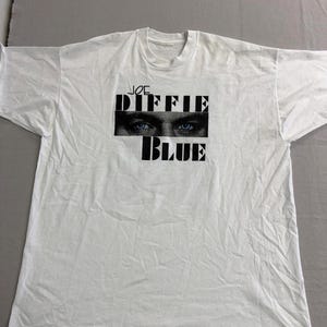 May include: White t-shirt featuring the text "JOE DIFFIE BLUE" in black block letters. The design includes a close-up of blue eyes. The shirt is made of a soft, likely cotton material.
