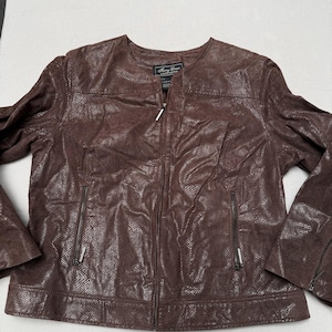Può includere: Una giacca marrone con un motivo testurizzato a pelle di serpente, caratterizzata da uno scollo rotondo, una chiusura frontale con zip e due tasche verticali con zip sul davanti. La giacca ha maniche lunghe con polsini dotati di cerniera.