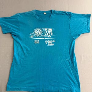 May include: A turquoise blue t-shirt with a white graphic that reads "RUN FOR LIFE" with a gear and other design elements. The shirt also features the Kentucky Fried Chicken logo and the Pepsi logo.