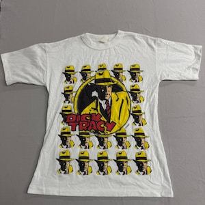 May include: White t-shirt featuring a graphic of Dick Tracy. The design includes a central image of the character in a yellow suit and hat, surrounded by smaller images of his face. The words "Dick Tracy" are printed in red and yellow.