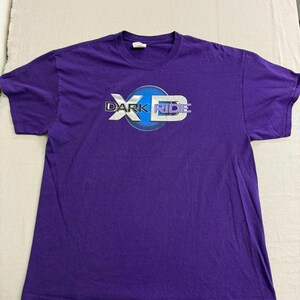 Könnte beinhalten: Ein lila T-Shirt mit dem Text "DARK RIDE" in einem weißen und blauen Design. Das Shirt ist aus einem weichen Material und hat einen Rundhalsausschnitt.