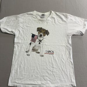 May include: A white short-sleeved t-shirt featuring a graphic of a brown and white dog holding a small American flag in its mouth. The text SPCA International is printed in black and red lettering to the right of the dog illustration.