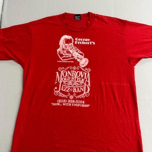 May include: A red t-shirt with a white graphic. The graphic includes the text "George Probert's" above an illustration of a musician playing a trumpet. Below is "Monrovia Old Style Jazz Band" and a phone number.