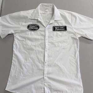 Peut inclure: Chemise blanche à manches courtes avec deux poches de poitrine. La poche gauche porte un écusson « COUNTY CORONER » et la poche droite un écusson « NIGHT SHIFT Supervisor ».