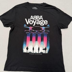 May include: A black short-sleeve t-shirt featuring the white text ABBA Voyage and London. The graphic displays four silhouettes of musicians on stage under bright pink and blue vertical light beams, with the text A CONCERT LIKE NO OTHER below.