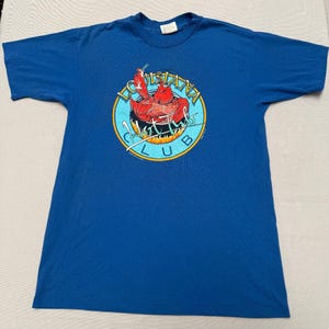 May include: A royal blue short-sleeve t-shirt featuring a circular graphic on the chest. The design depicts a red crawfish inside a black pot with flames, surrounded by the text Louisiana Hot Tub Club in yellow and white lettering.