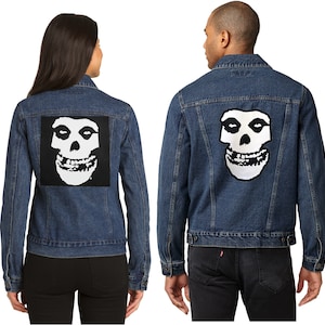 May include: Two denim jackets, one on a woman and one on a man, each with a large, square black patch with a white skull design on the back. The jackets are a medium blue wash and have classic denim jacket details.