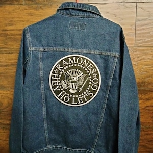 May include: A blue denim jacket with a black Ramones patch on the back. The patch features a presidential seal with an eagle and the words "Hey Ho Let's Go".