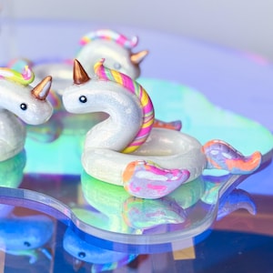 Peut inclure: Une collection de petites figurines de licornes blanc irisé, chacune dotée d'une corne en cuivre métallique et d'une crinière et d'une queue multicolores vives. Les licornes ont la forme de bouées, avec des ailes et des yeux noirs.
