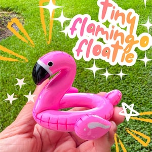 Peut inclure: Une petite bouée gonflable rose vif en forme de flamant rose avec un bec noir et des détails blancs sur le visage. Le texte "Tiny flamingo floatie" est écrit dans une police blanche et orange stylisée au-dessus de l'objet, avec des étoiles.