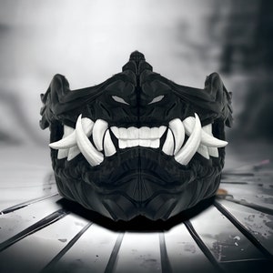 May include: A black face mask featuring a sculpted, textured surface with prominent, sharp white fangs and teeth. The design includes a central ridge and angular contours, creating a fierce, detailed appearance that covers the nose and mouth area.