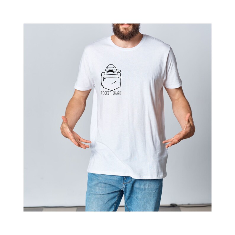 May include: White t-shirt with a black and white illustration of a shark in a pocket. The text "Pocket Shark" is printed below the illustration.