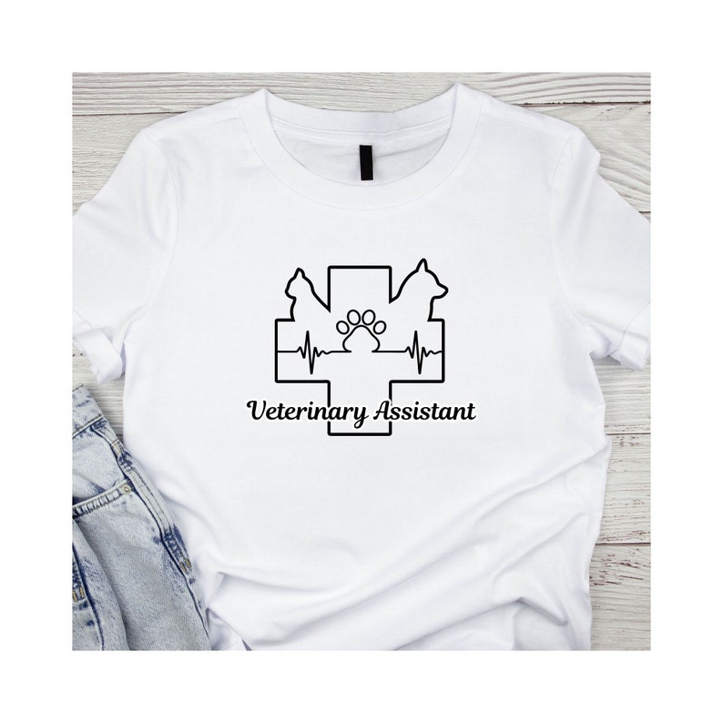 Peut inclure: T-shirt blanc avec un motif graphique noir repr&eacute;sentant une croix m&eacute;dicale avec une empreinte de patte et une ligne de battement de c&oelig;ur. Le texte "Veterinary Assistant" est situ&eacute; sous le graphique.