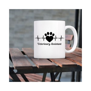 Peut inclure: Une tasse &agrave; caf&eacute; en c&eacute;ramique blanche avec un imprim&eacute; de patte noire et un c&oelig;ur graphique, et le texte "Assistant v&eacute;t&eacute;rinaire".