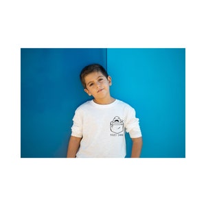 May include: A young person wearing a white long-sleeved t-shirt with a black and white pocket shark graphic and the text "Pocket Shark". The person is standing in front of a blue wall.