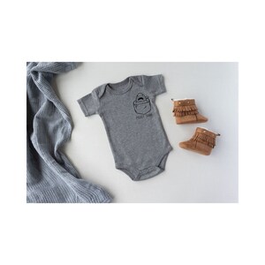 May include: A gray baby onesie with a pocket shark graphic and the text "Pocket Shark". The onesie is laying on a white surface with a gray blanket and brown baby booties.