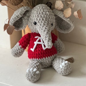 Könnte beinhalten: Ein grauer gehäkelter Elefant, der einen roten Pullover mit einem weißen Buchstaben 'A' auf der Vorderseite trägt.