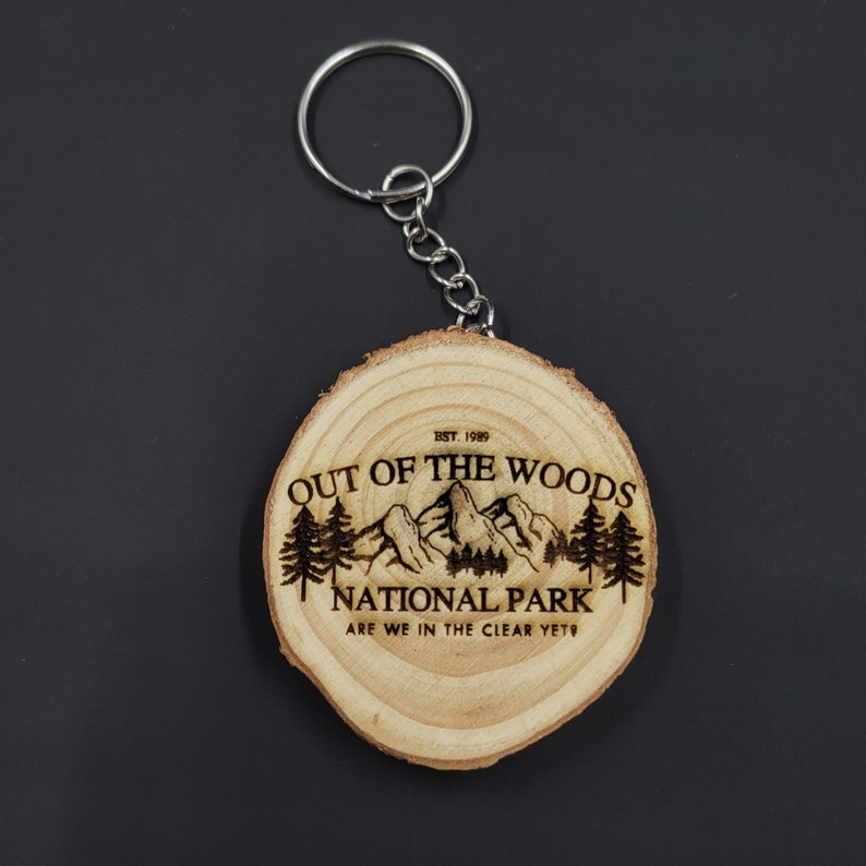 May include: A wooden keychain with a burnt design of a mountain range and trees. The text reads "EST. 1989 OUT OF THE WOODS NATIONAL PARK ARE WE IN THE CLEAR YET".