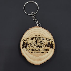 May include: A wooden keychain with a laser-engraved design of a mountain range and trees. The text "EST. 1989 OUT OF THE WOODS NATIONAL PARK ARE WE IN THE CLEAR YET?" is engraved on the keychain.