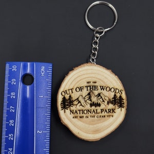 May include: A wooden keychain with a brown wood grain finish. The keychain features a laser-engraved design of a mountain range with trees and the text "Out of the Woods National Park. Est. 1989. Are we in the clear yet?"