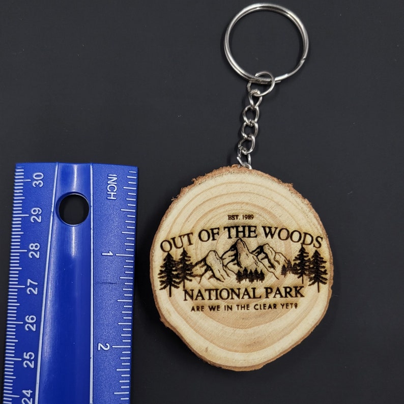 May include: A wooden keychain with a brown wood grain finish. The keychain features a laser-engraved design of a mountain range with trees and the text "Out of the Woods National Park. Est. 1989. Are we in the clear yet?"