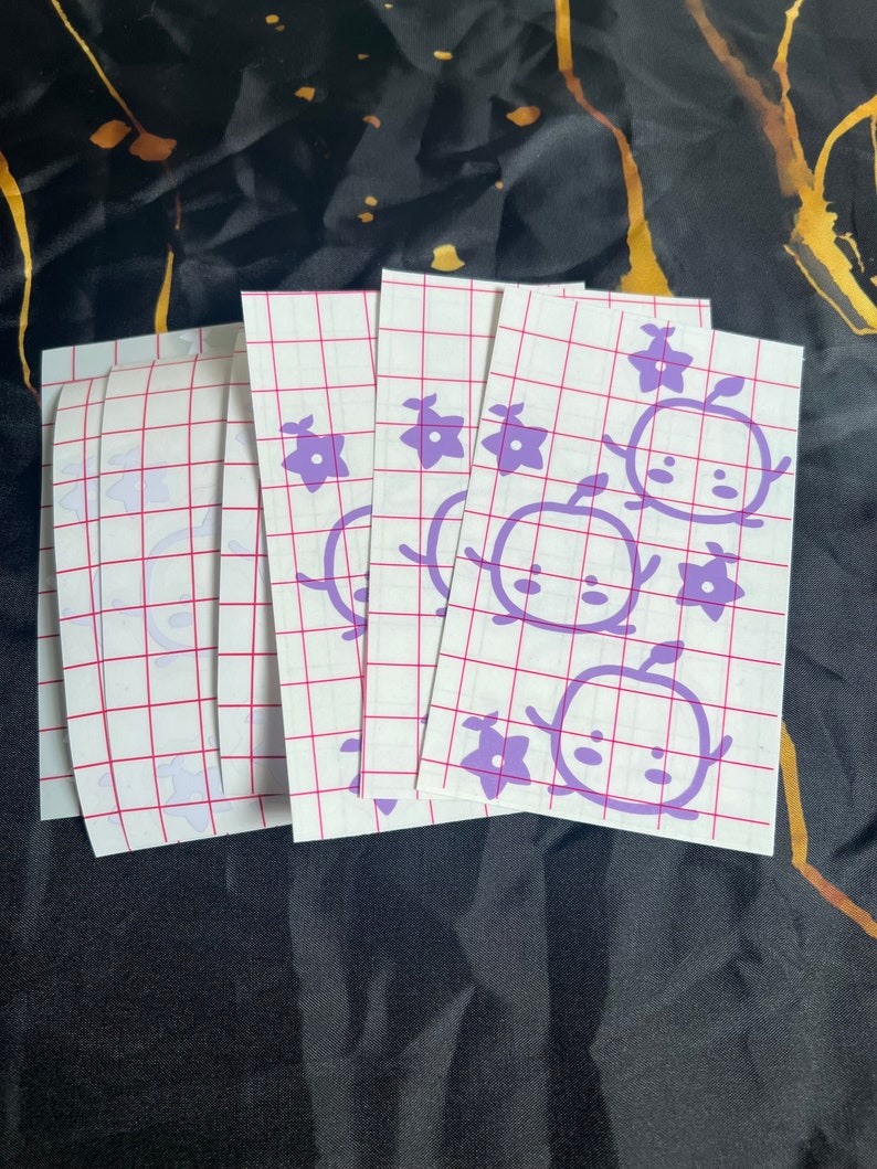 May include: A set of five purple vinyl stickers featuring a cartoon character with a flower and an apple. The stickers are on a white sheet with a grid pattern.