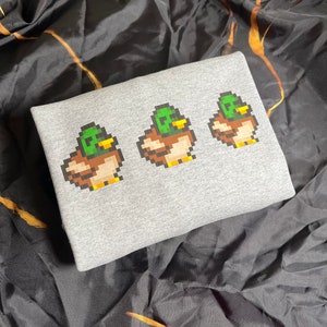 Pu&ograve; includere: Felpa grigia con un design pixelato di tre anatre in fila. Le anatre sono verdi, marroni e gialle.