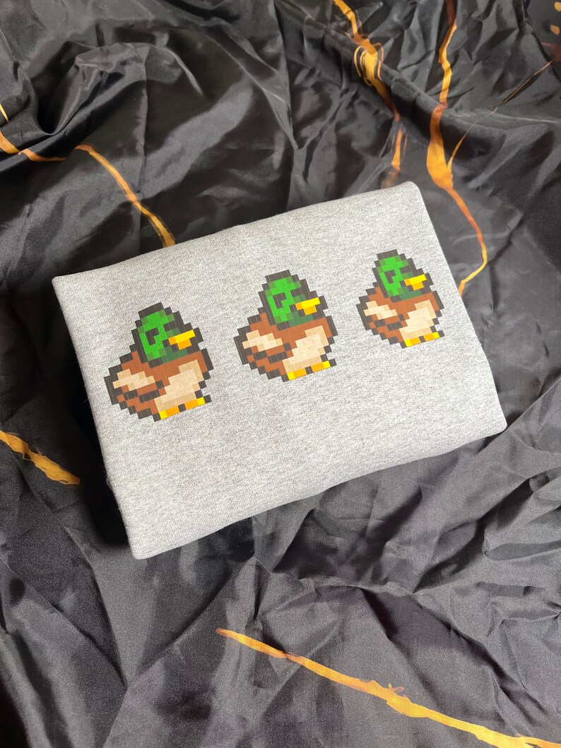 Pu&ograve; includere: Felpa grigia con un design pixelato di tre anatre in fila. Le anatre sono verdi, marroni e gialle.