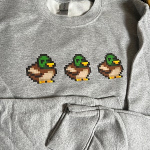 Pu&ograve; includere: Una felpa grigia con un design a pixel di tre anatre in fila. Le anatre sono verdi, marroni e gialle.