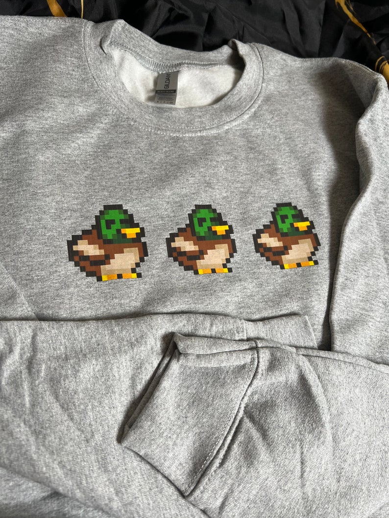 Pu&ograve; includere: Una felpa grigia con un design a pixel di tre anatre in fila. Le anatre sono verdi, marroni e gialle.