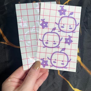 May include: Two sheets of clear transfer paper with purple vinyl cutouts of cartoon characters with flowers. The characters have round bodies, small arms, and large eyes. The flowers are five-petaled.