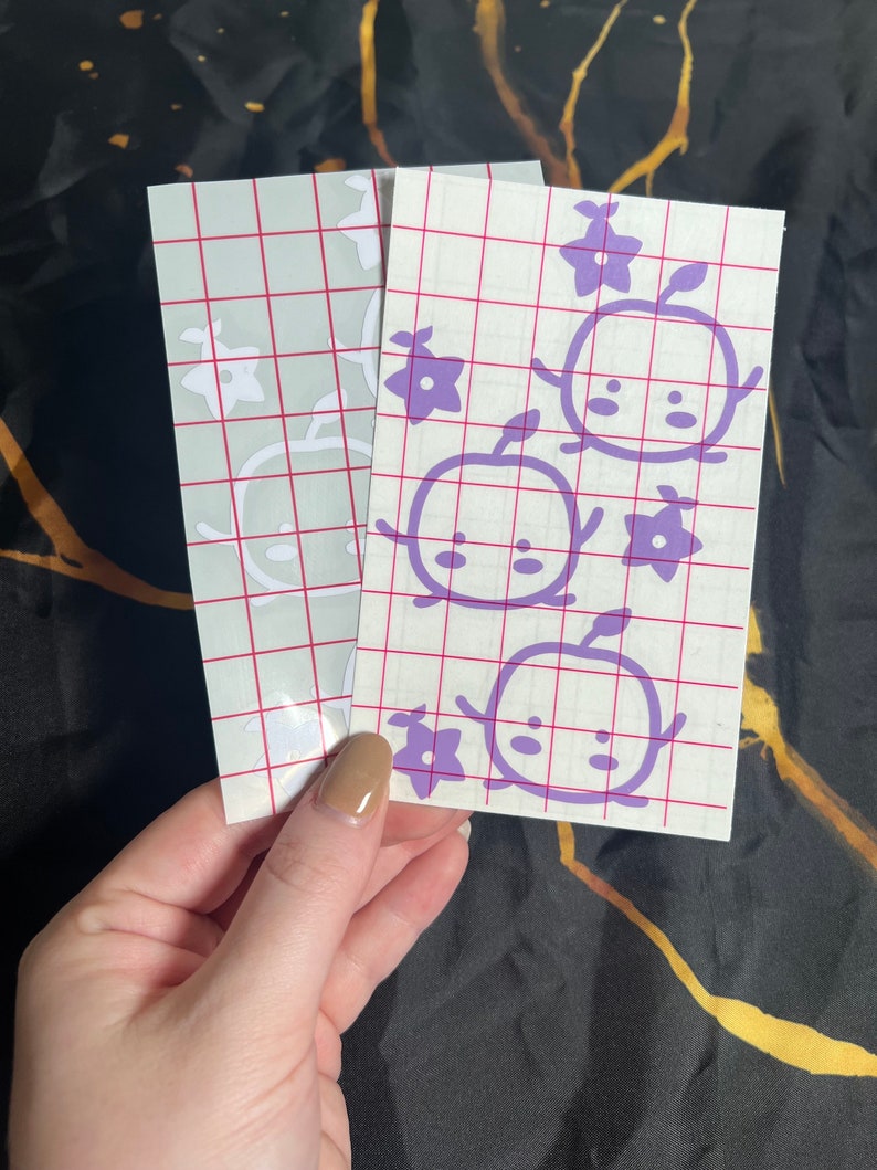 May include: Two sheets of clear transfer paper with purple vinyl cutouts of cartoon characters with flowers. The characters have round bodies, small arms, and large eyes. The flowers are five-petaled.