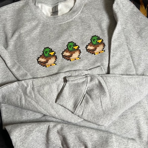 Pu&ograve; includere: Una felpa grigia con un design pixelato di tre anatre che indossano cappelli. Le anatre sono in fila, rivolte verso il lato destro dell'immagine.