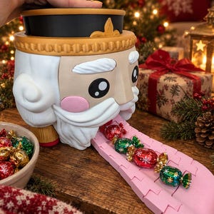May include: A nutcracker-shaped candy dispenser with a tan face, white beard, and black hat. It features a pink tongue-shaped slide extending from its mouth, holding several red and green foil-wrapped candies in a line on the surface.