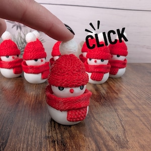 Può includere: Una collezione di piccole statuette a forma di pupazzo di neve. Ogni pupazzo di neve ha un corpo bianco, un cappello rosso lavorato a maglia con un pompon bianco e una sciarpa rossa. La parola "CLICK" è scritta in nero, con una mano che indica il pupazzo di neve.